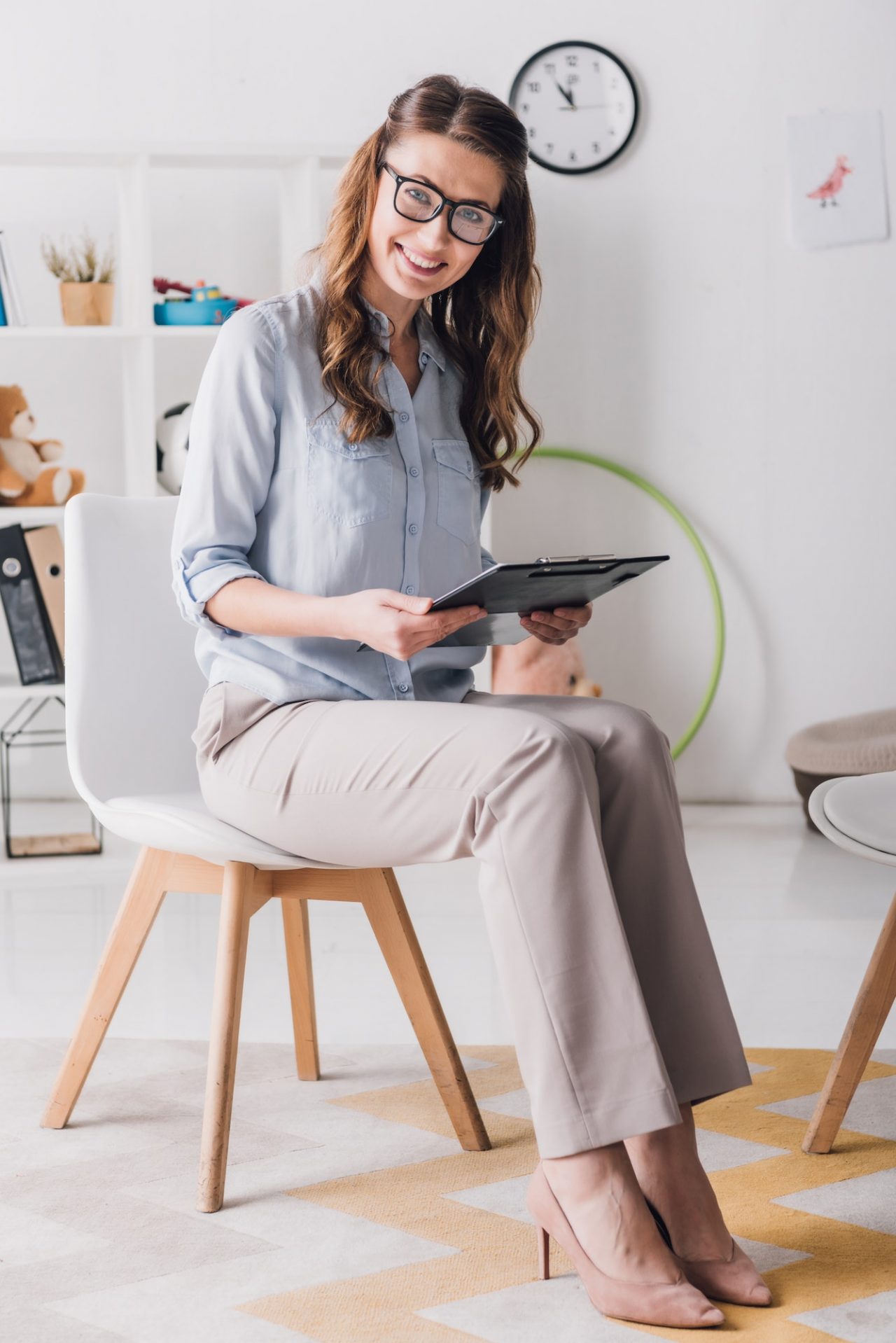 smiling-child-psychologist-with-clipboard-sitting-on-chair-in-office-and-looking-at-camera.jpg